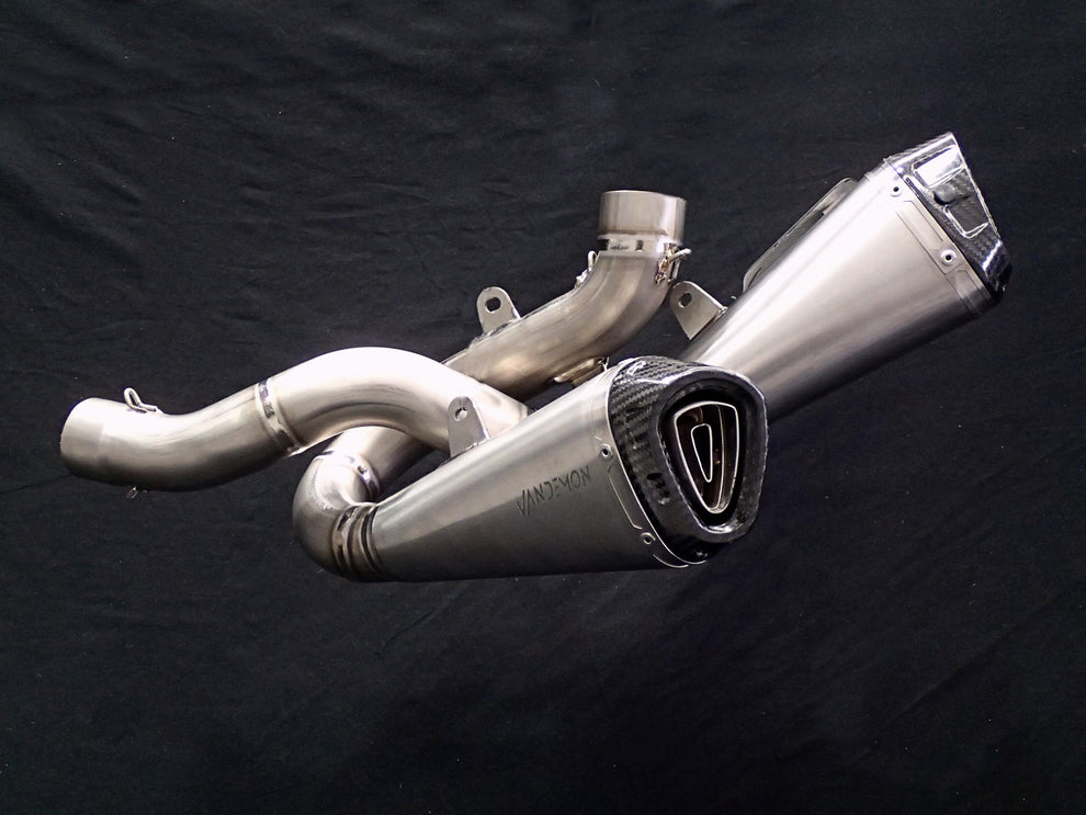 A polished stainless steel motorcycle exhaust muffler with a black and silver triangular badge, shown against a black fabric background.